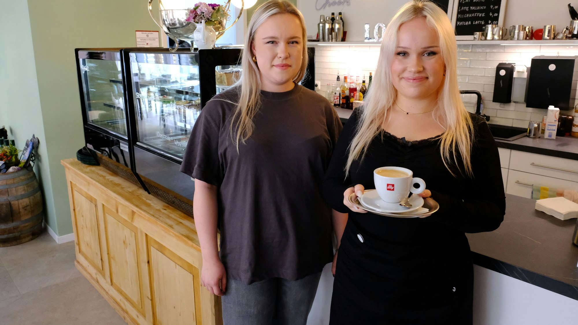 Hanna Schnigge und ihre Schwester Leni im Gastraum der Café-Bar. Leni hält eine Tasse Kaffee in der Hand.