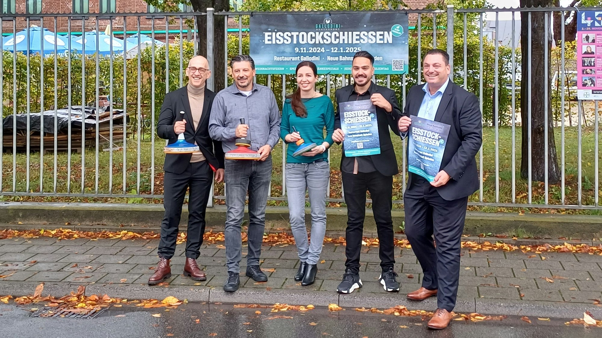 Vier Männer und eine Frau stehen mit Eisstöcken vor einem Plakat.