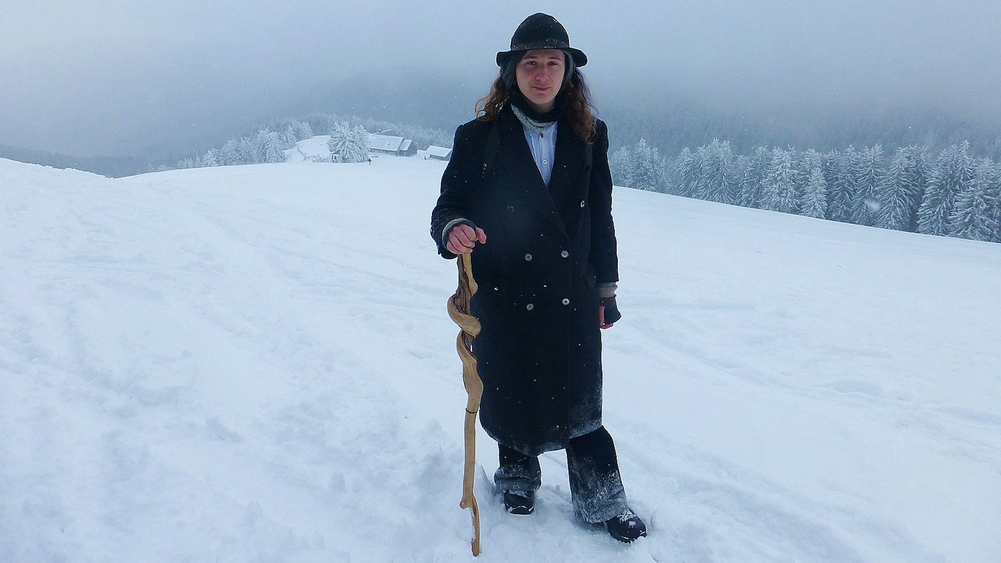 Ein Mann in Mantel stampft mit Wanderstock durch Schnee. Im Hintergrund befindet sich ein verschneiter Wald.