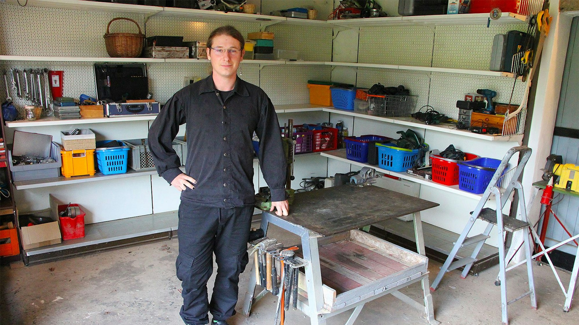 Ein junger Mann mit Brille lehnt sich auf einen Tisch in einem Raum mit Werkzeug.