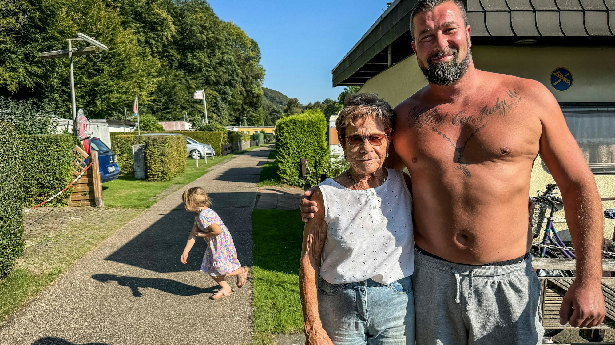 Trudi Decker und Patrick Blatzheim stehen Arm in Arm auf einem Campingplatz, er trägt kein T-Shirt.