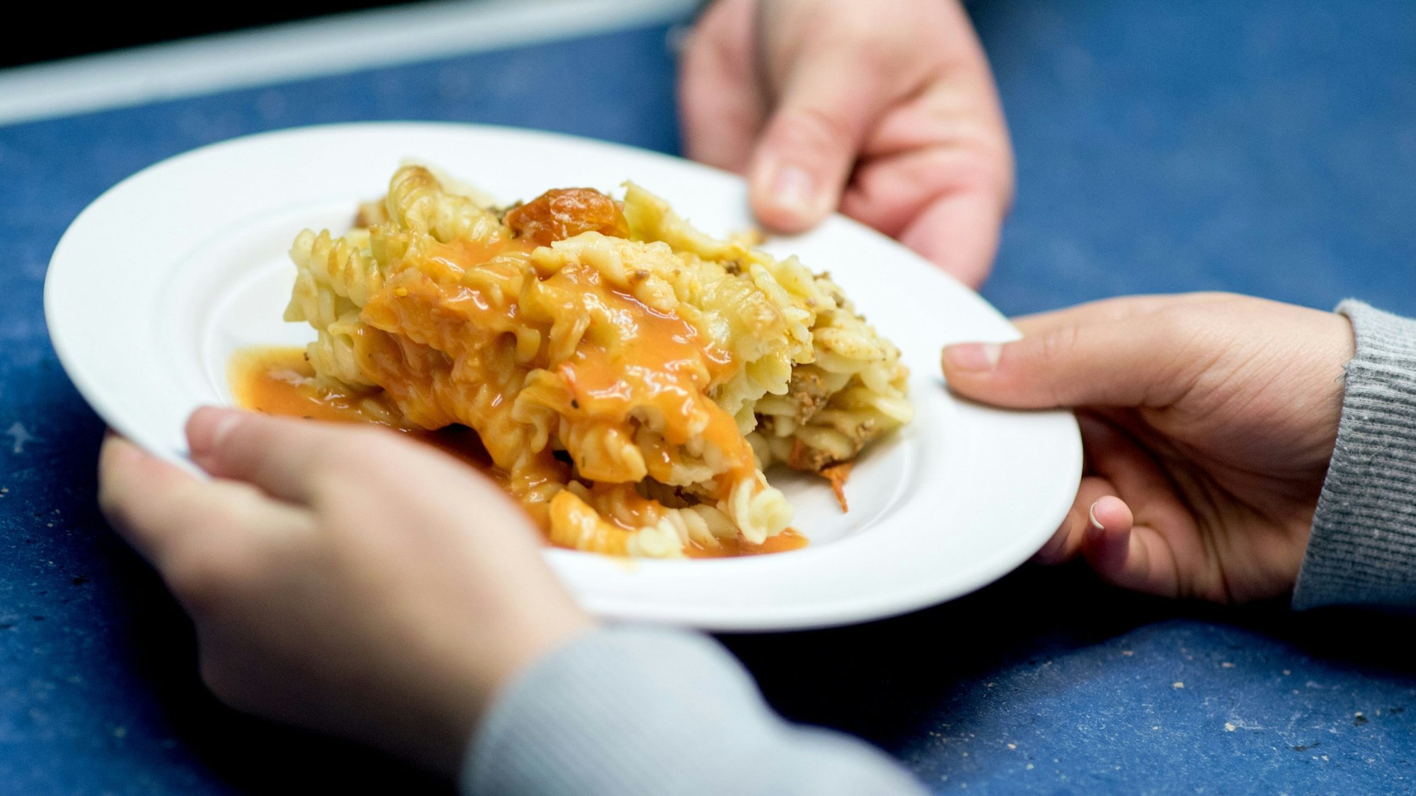 Ein Teller, gefüllt mit Nudelauflauf und Soße, wandert aus der Hand einer Mensa-Mitarbeiterin in die Hand eines Schülers.
