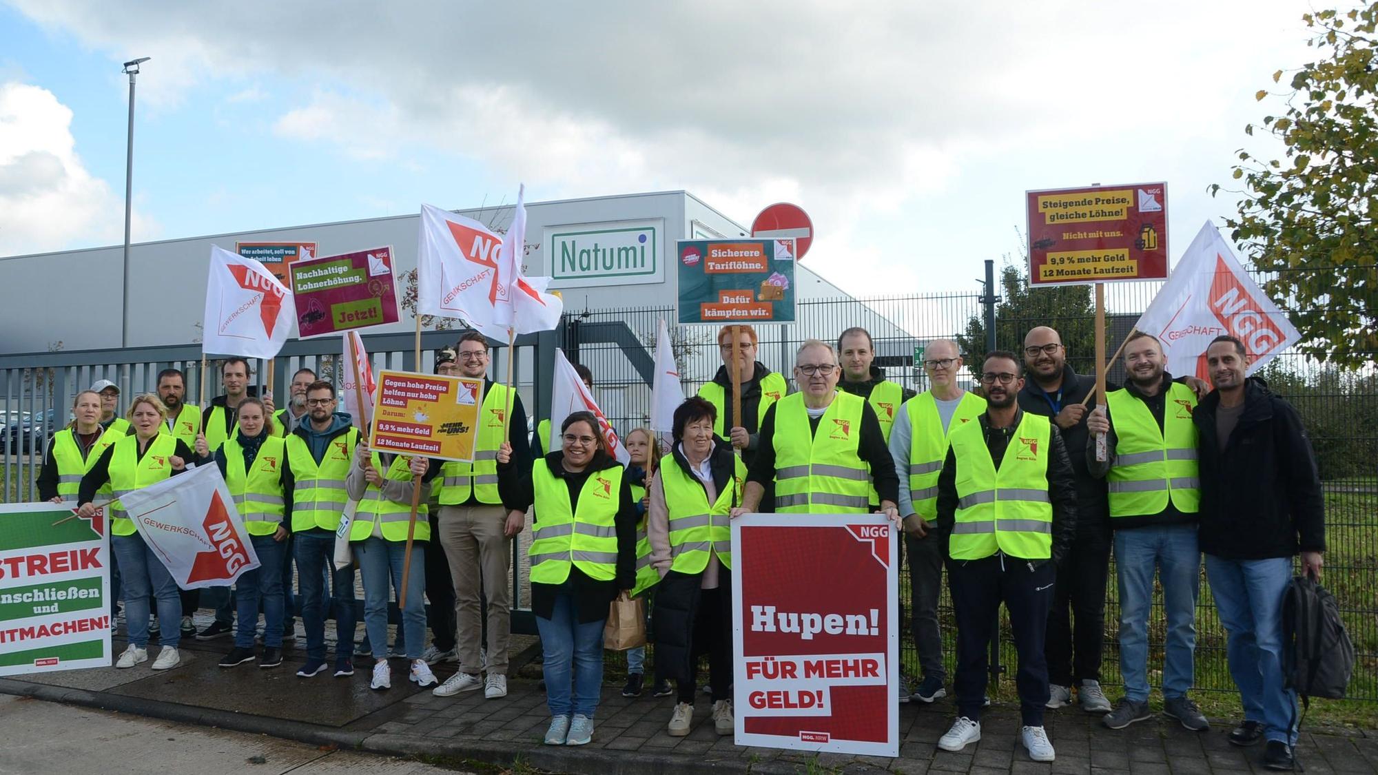 Männer und Frauen in gelben Warnwesten stehen vor einem Firmengelände. Sie tragen Plakate mit Aufforderungen wie "Hupen für mehr Geld" oder "Streik - anschließen und mitmachen."