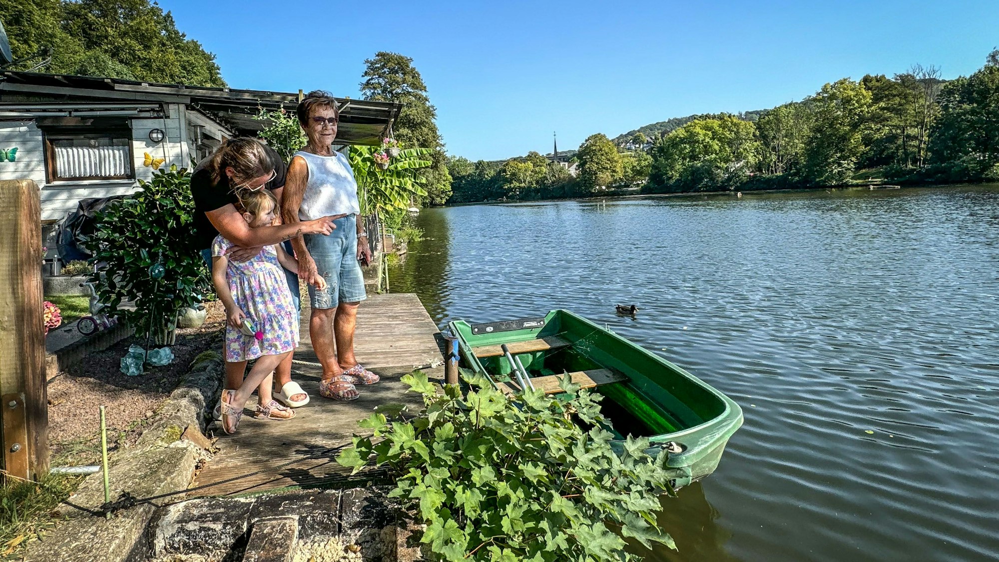 An der Agger in Loope schaut Trudi Decker gemeinsam mit Denise Kurth und deren Tochter Amelie aufs Wasser. Vor ihnen ist ein kleines, grünes Boot zu sehen. Der Himmel ist blau, die Sonne scheint.