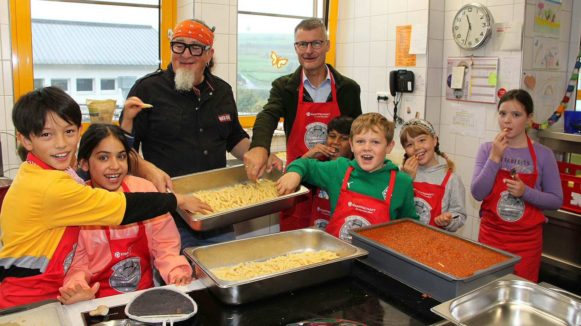 Zwei Männer und eine Gruppe Kinder stehen in einer Küche, in der große Behälter mit Nudeln und einer roten Sauce stehen.