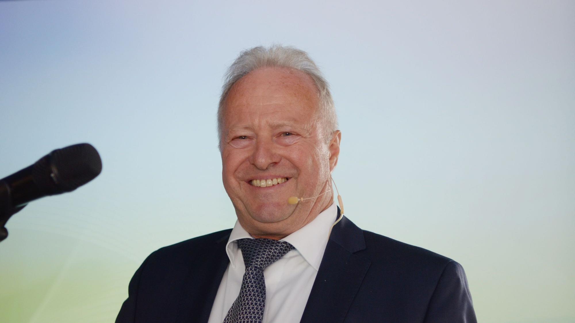 Michael Lauster, vor sich ein Mikrofon, lacht in die Kamera.