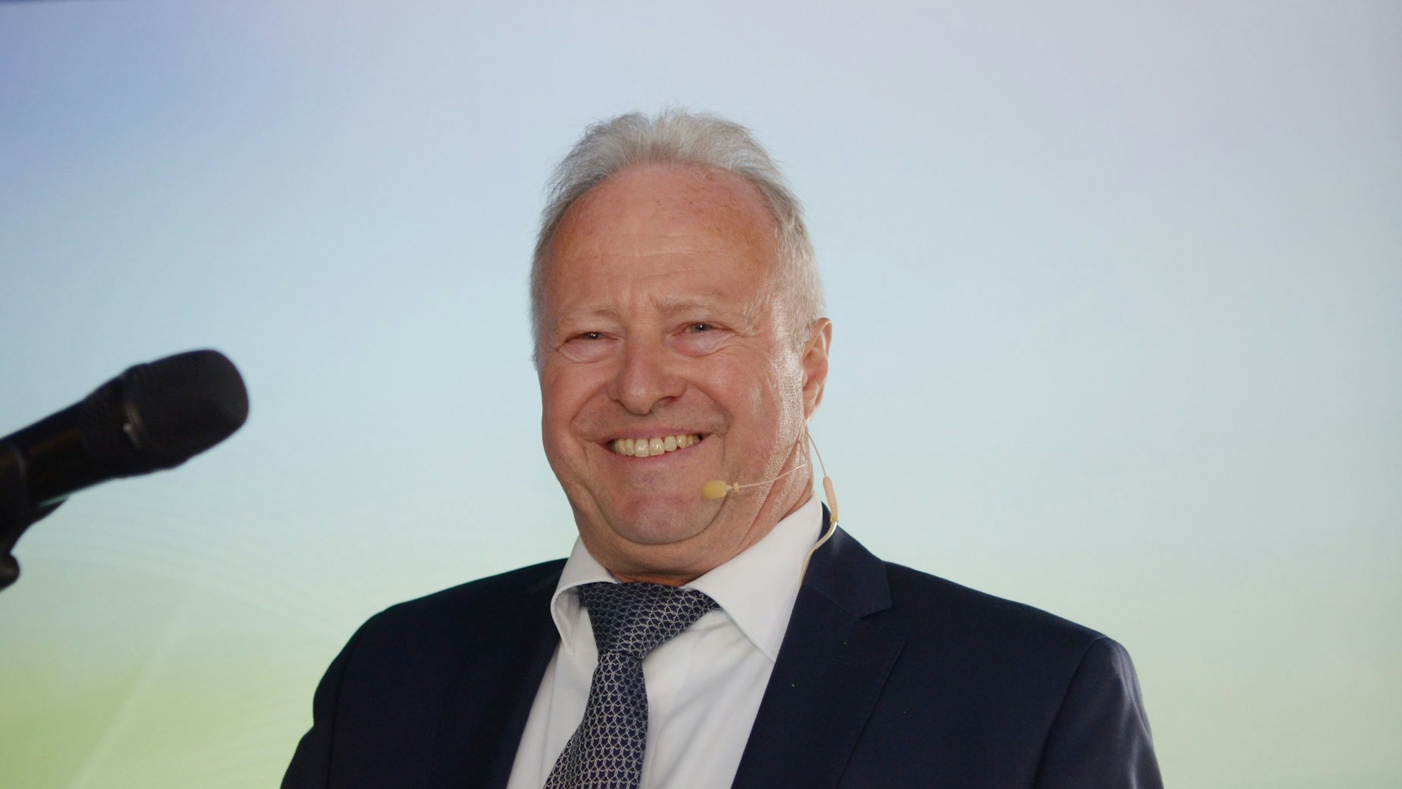 Michael Lauster, vor sich ein Mikrofon, lacht in die Kamera.