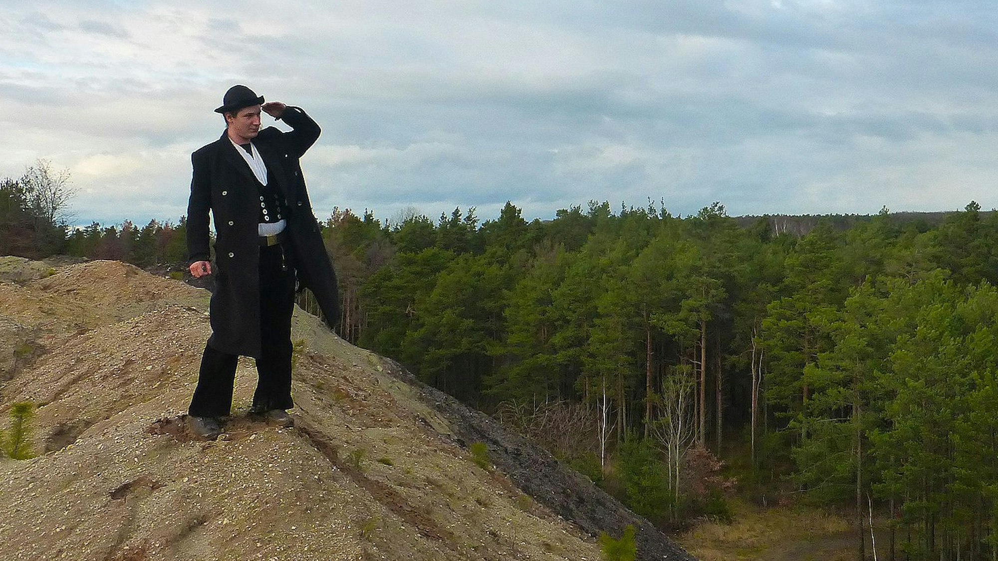 Ein Mann in traditioneller Bekleidung schaut auf einem Hügel in die Ferne. Im Hintergrund befindet sich ein Wald.