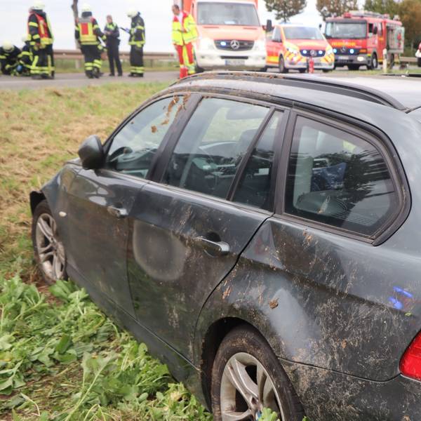 Ein lädierter Wagen mit Beulen und verdreckter Seite steht in einem Feld. Im Hintergrund stehen Feuerwehr und Rettungskräfte mit Fahrzeugen.