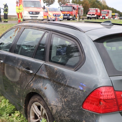 Ein lädierter Wagen mit Beulen und verdreckter Seite steht in einem Feld. Im Hintergrund stehen Feuerwehr und Rettungskräfte mit Fahrzeugen.