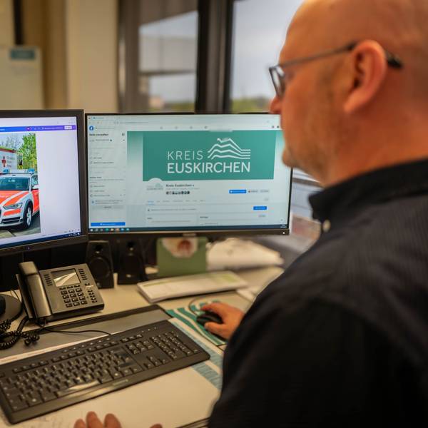 Ein Mann im schwarzen Hemd sitzt vor zwei Computerbildschirmen.