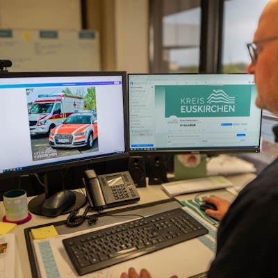 Ein Mann im schwarzen Hemd sitzt vor zwei Computerbildschirmen.