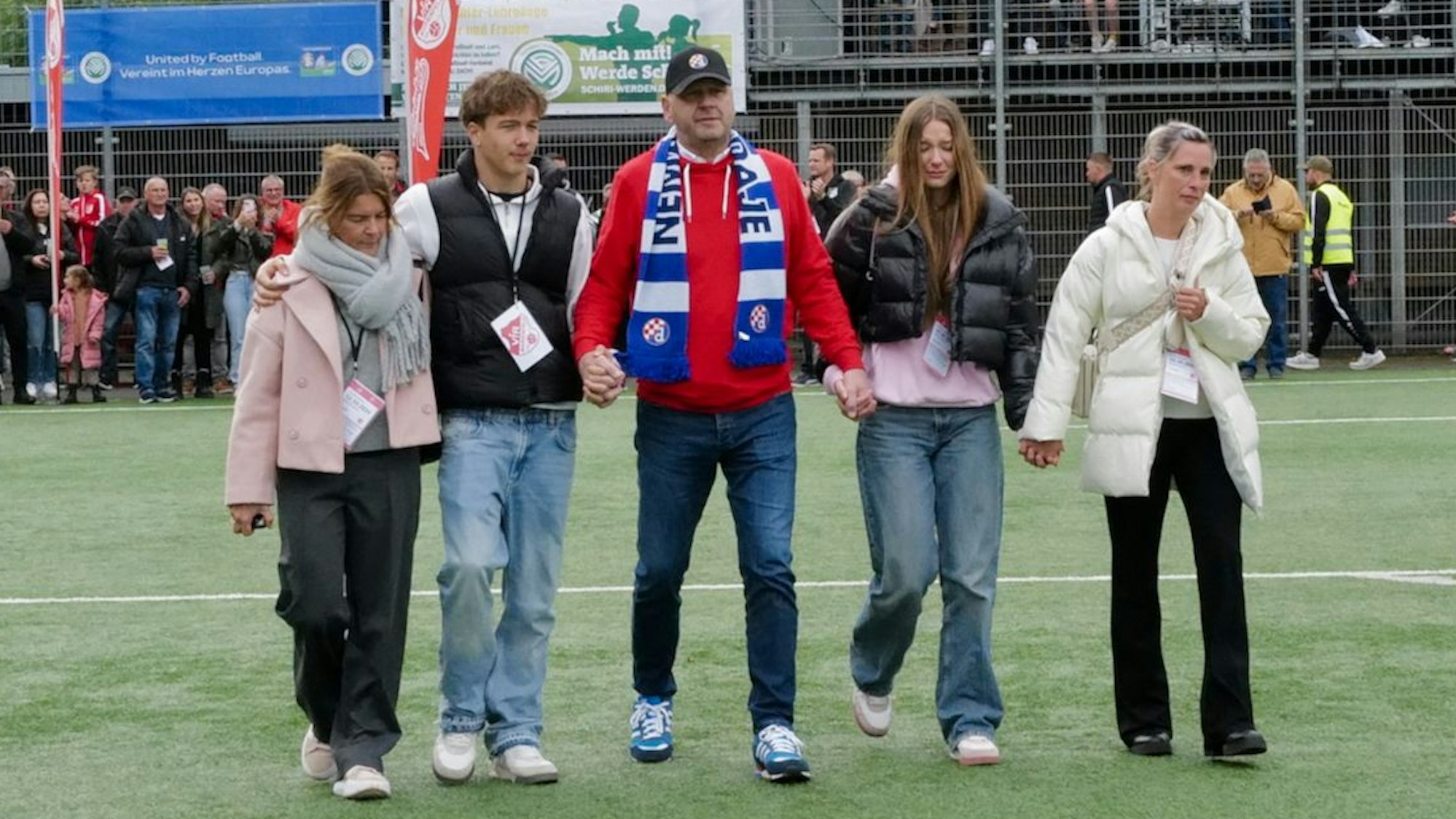 Fünf Personen laufen Hand in Hand auf ein Fußballspielfeld.