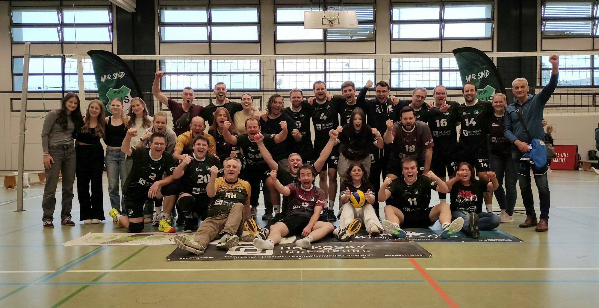 Eine Mannschaft in Sportkleidung hat sich zum Gruppenfoto aufgestellt, im Hintergrund steht ein Volleyballnetz.