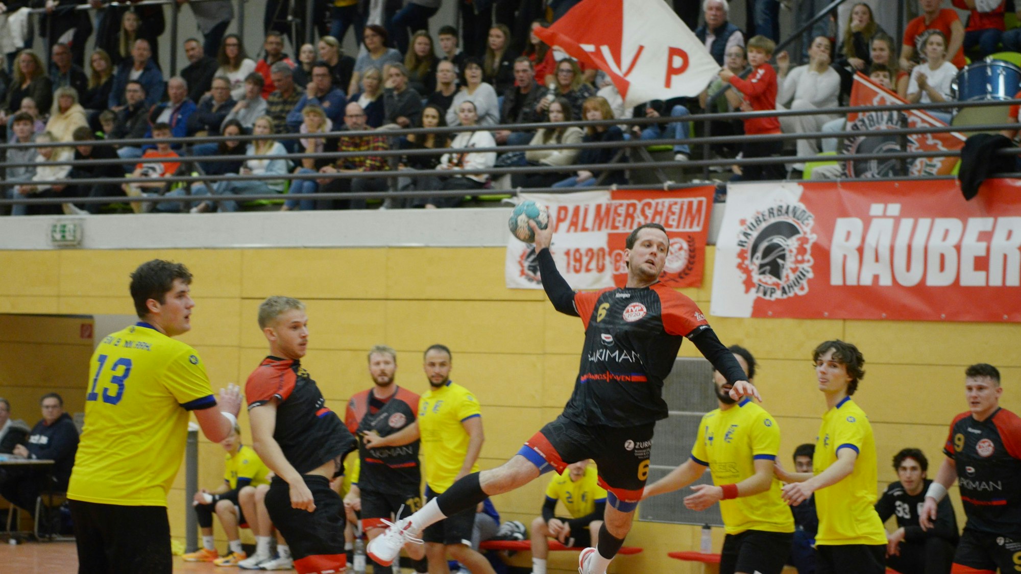 Fiedler beim Sprungwurf – Mitspieler und Gegner schauen zu. Im Hintergrund ein voll besetzter Teil der Tribüne.