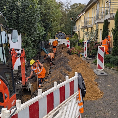 Das Foto zeigt einen Bagger vor einer Baugrube, in der Bauarbeiter gerade Glasfaserkabel verlegen.