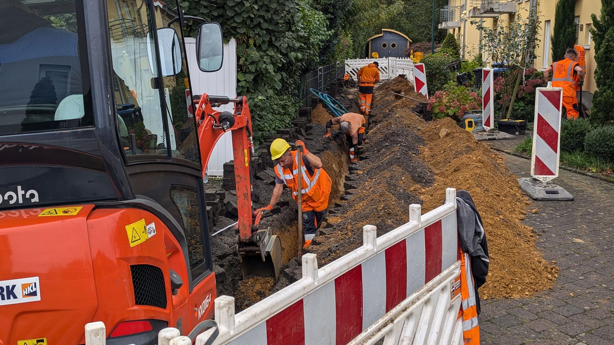 Das Foto zeigt einen Bagger vor einer Baugrube, in der Bauarbeiter gerade Glasfaserkabel verlegen.