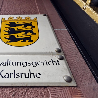 Ein Hinweisschild mit dem Landeswappen von Baden-Württemberg und dem Schriftzug ·Verwaltungsgericht Karlsruhe·, aufgenommen vor dem Gericht.