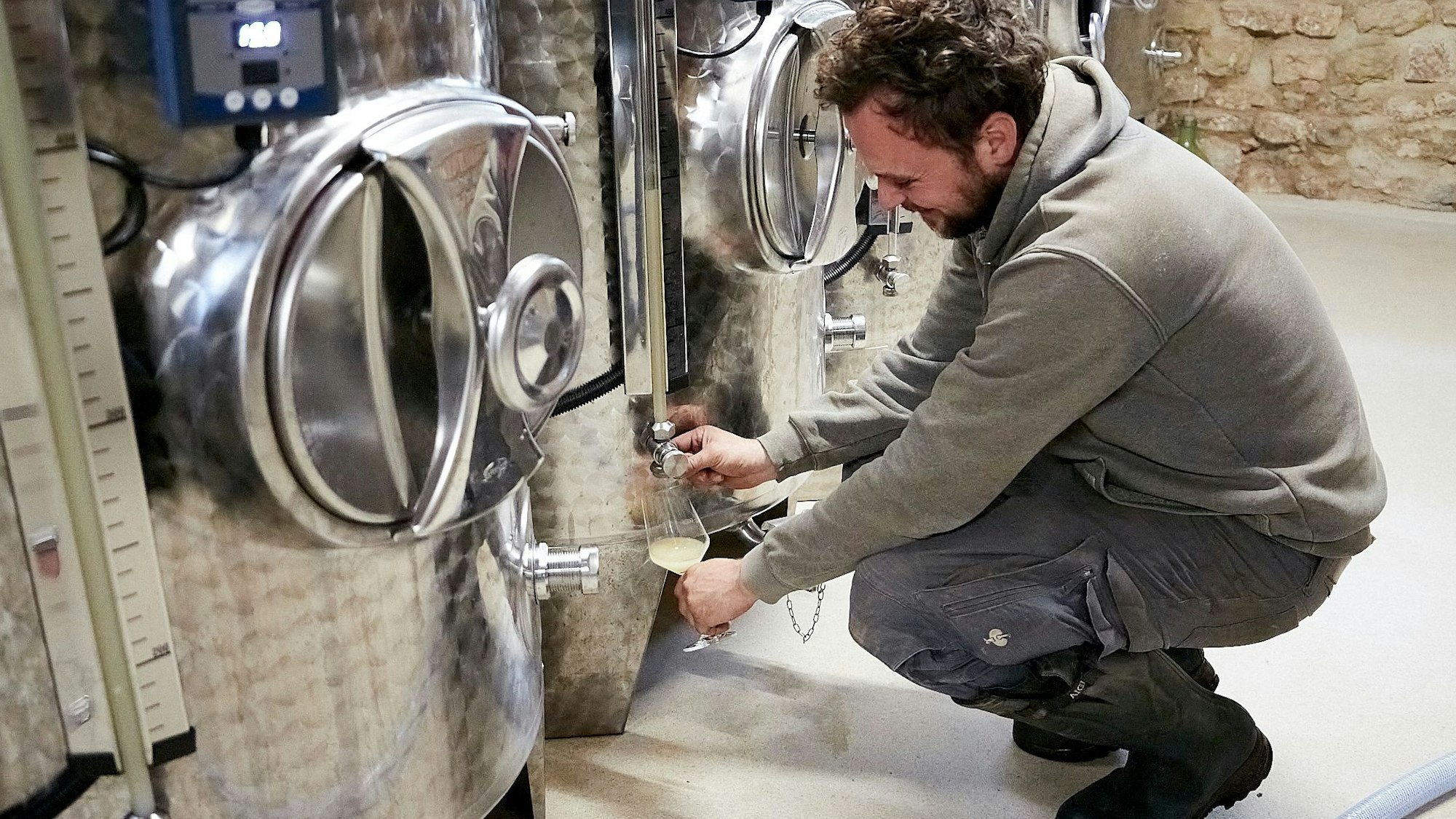 Markus Schäfer hockt vor einem silberfarbenen Edelstahltank und füllt eine helle, trübe Flüssigkeit in ein Weinglas.