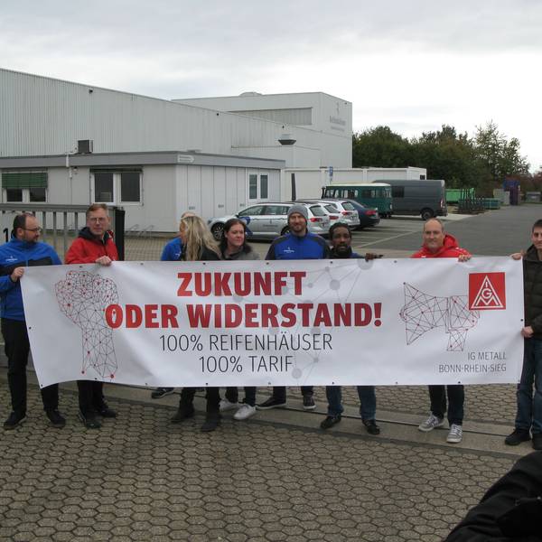 Acht Männer und zwei Frauen vor einem Werkstor mit einem Protest-Transparent der IG Metall. Rechts im Vordergrund steht ein Fernseh-Kameramann.