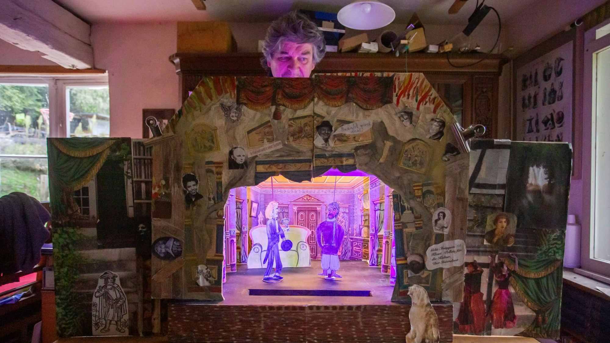 Ein Mann steht hinter einem Papiertheater, das eine Szene einer Komödie zeigt.