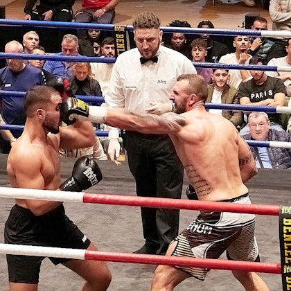 Zwei Boxer kämpfen unter der Aufsicht eines Ringrichters. Im Hintergrund verfolgen zahlreiche Zuschauer den Kampf.