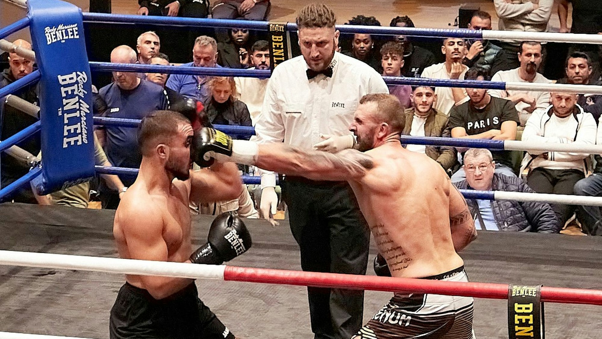 Zwei Boxer kämpfen unter der Aufsicht eines Ringrichters. Im Hintergrund verfolgen zahlreiche Zuschauer den Kampf.