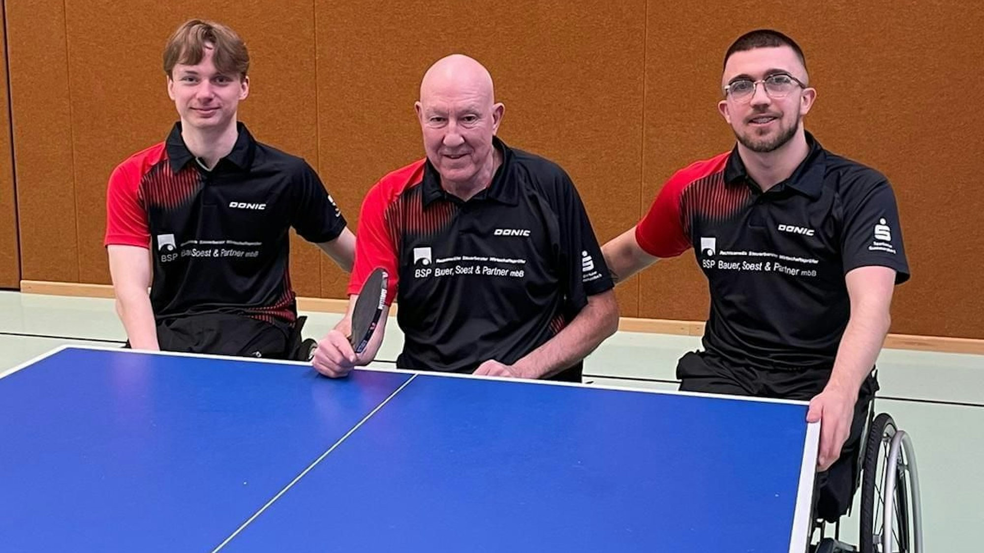 Ein Rollstuhl-Tischtennis-Trio an einer Tischtennisplatte.