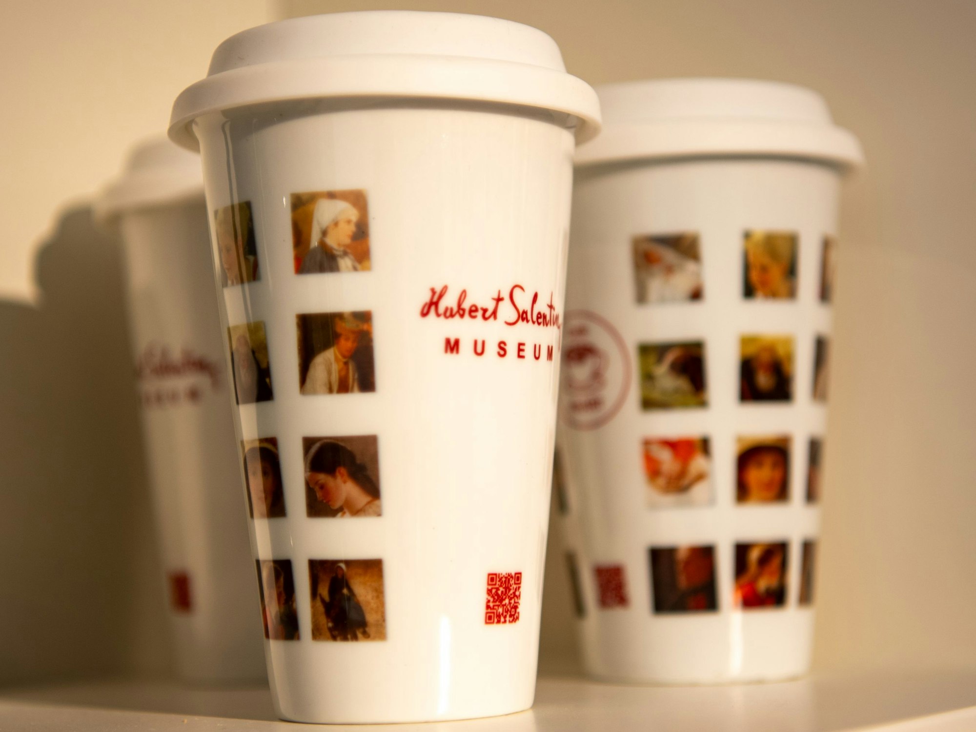 Mehrweg-Kaffeebecher mit dem Aufdruck „Hubert Salentin Museum“ und verschiedenen Bildausschnitten von Salentins Gemälden stehen in einem Regal.