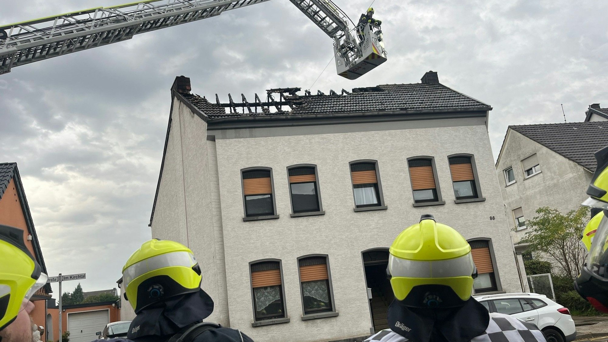 Feuerwehrleute stehen vor einem Mehrfamilienhaus, über dem ausgebrannten Dach schwebt der Korb der Drehleiter.
