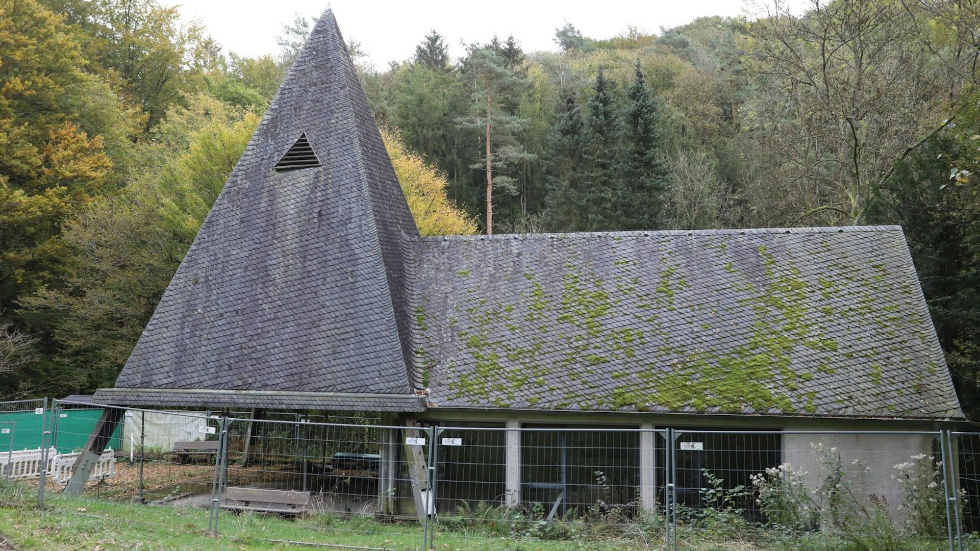 Ein Bauzaun umgibt eine Kapelle, auf deren Dach zum Teil grünes Moos wächst.