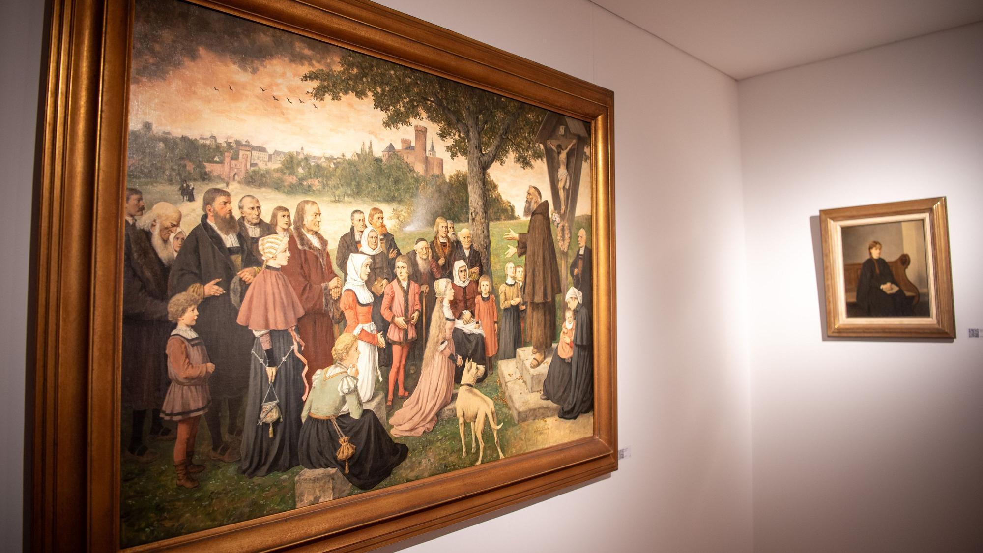 Das Bild „Die Kapuzinerpredigt“ von Hubert Salentin zeigt eine Gruppe Menschen, die sich außerhalb einer Stadt auf einem Hügel an einem Kreuz versammelt hat.