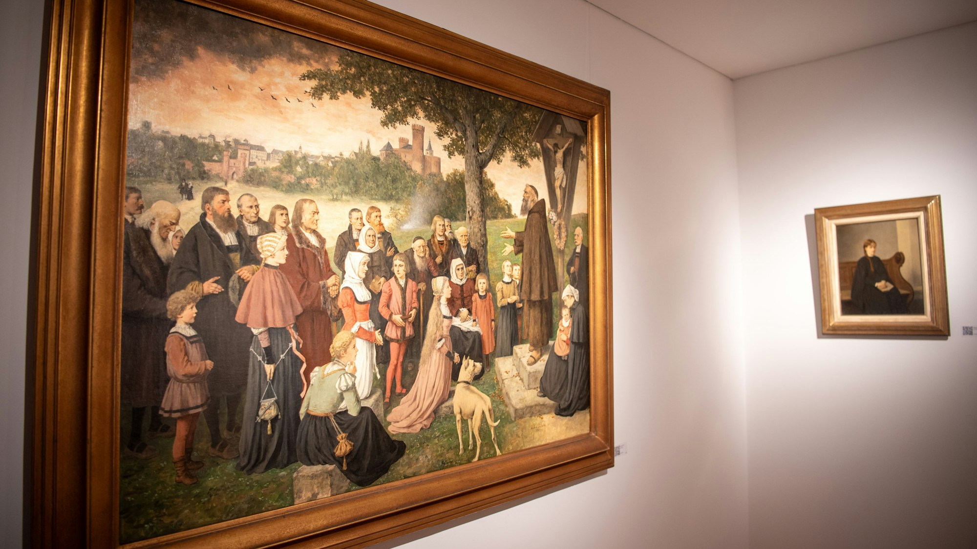 Das Bild „Die Kapuzinerpredigt“ von Hubert Salentin zeigt eine Gruppe Menschen, die sich außerhalb einer Stadt auf einem Hügel an einem Kreuz versammelt hat.