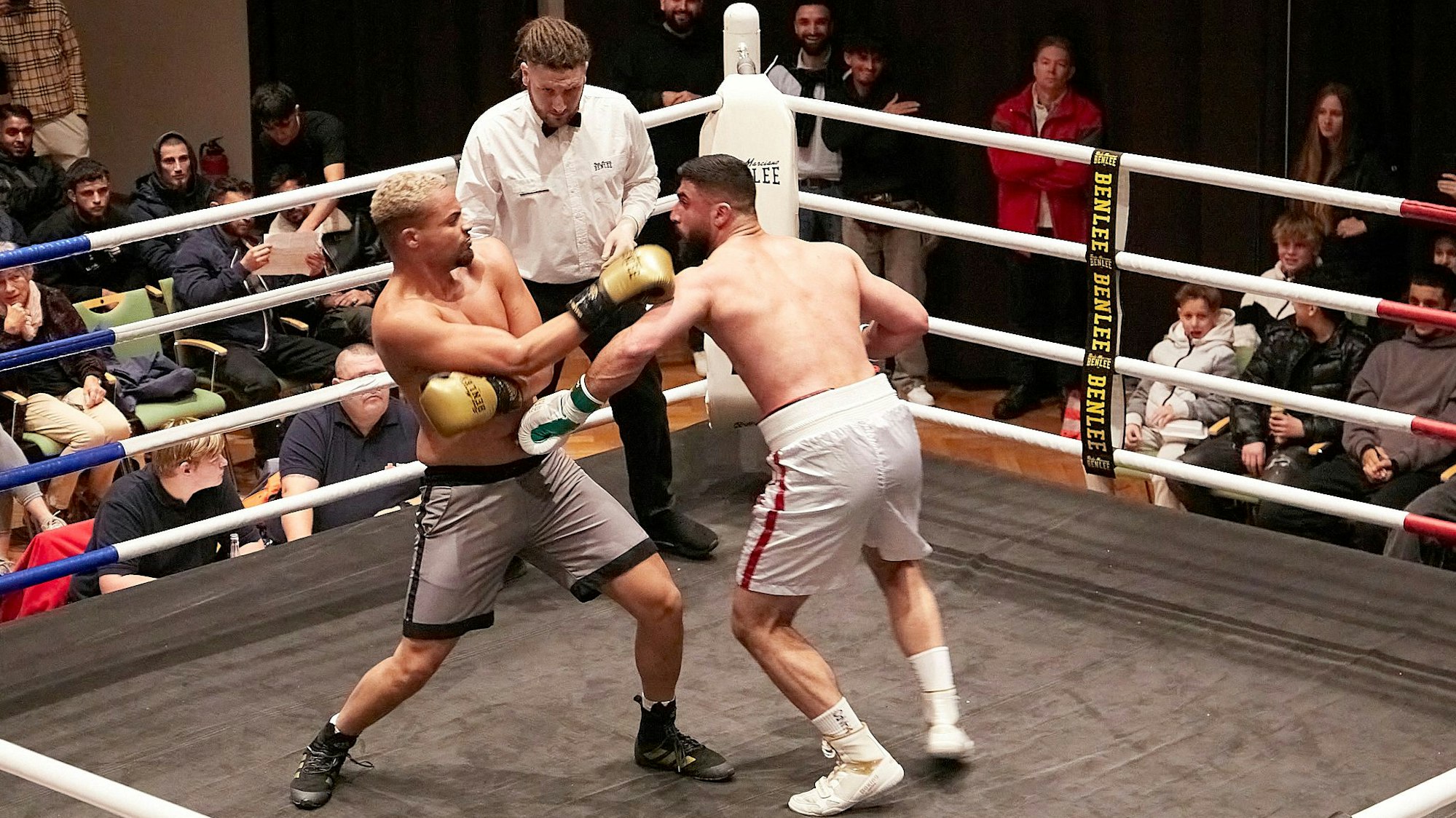 Zwei Boxer kämpfen unter der Aufsicht eines Ringrichters. Im Hintergrund verfolgen zahlreiche Zuschauer den Kampf.