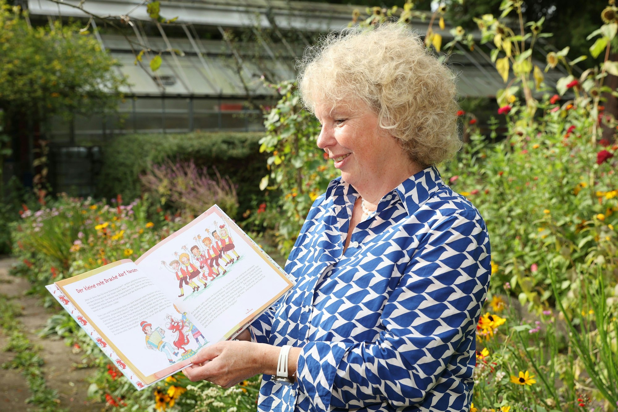 Brigitte Hintzen-Bohlen hält ihr neues Buch aufgeschlagen in der Hand, zu sehen sind Zeichnungen von einem kleinen roten Drachen in einer Tanzgruppe.