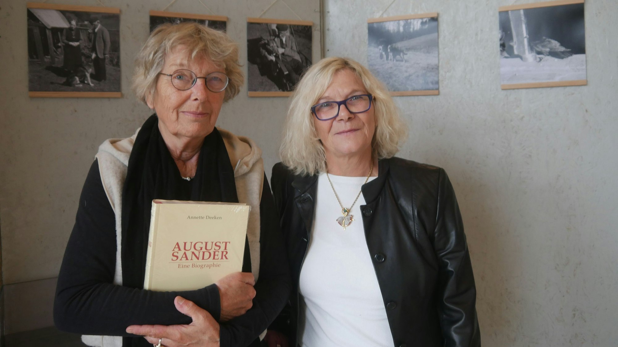 Zwei Frauen stehen vor alten Schwarz-Weiß-Fotos, eine hält ein Buch im Arm.