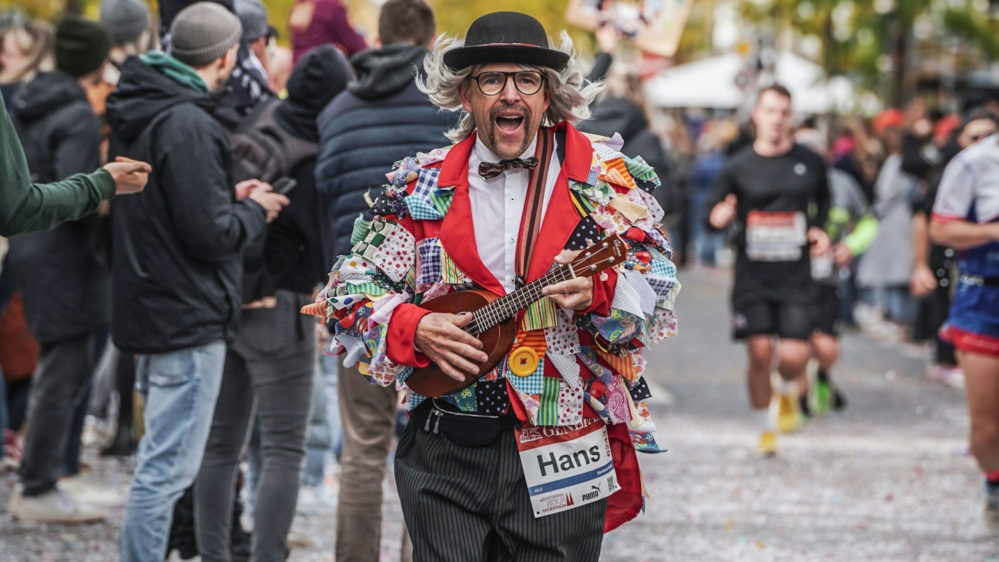 Ein Mann läuft im Kostüm von Hans Süper, mit Ukulele und singend. Hinter ihm laufen andere Sportler.
