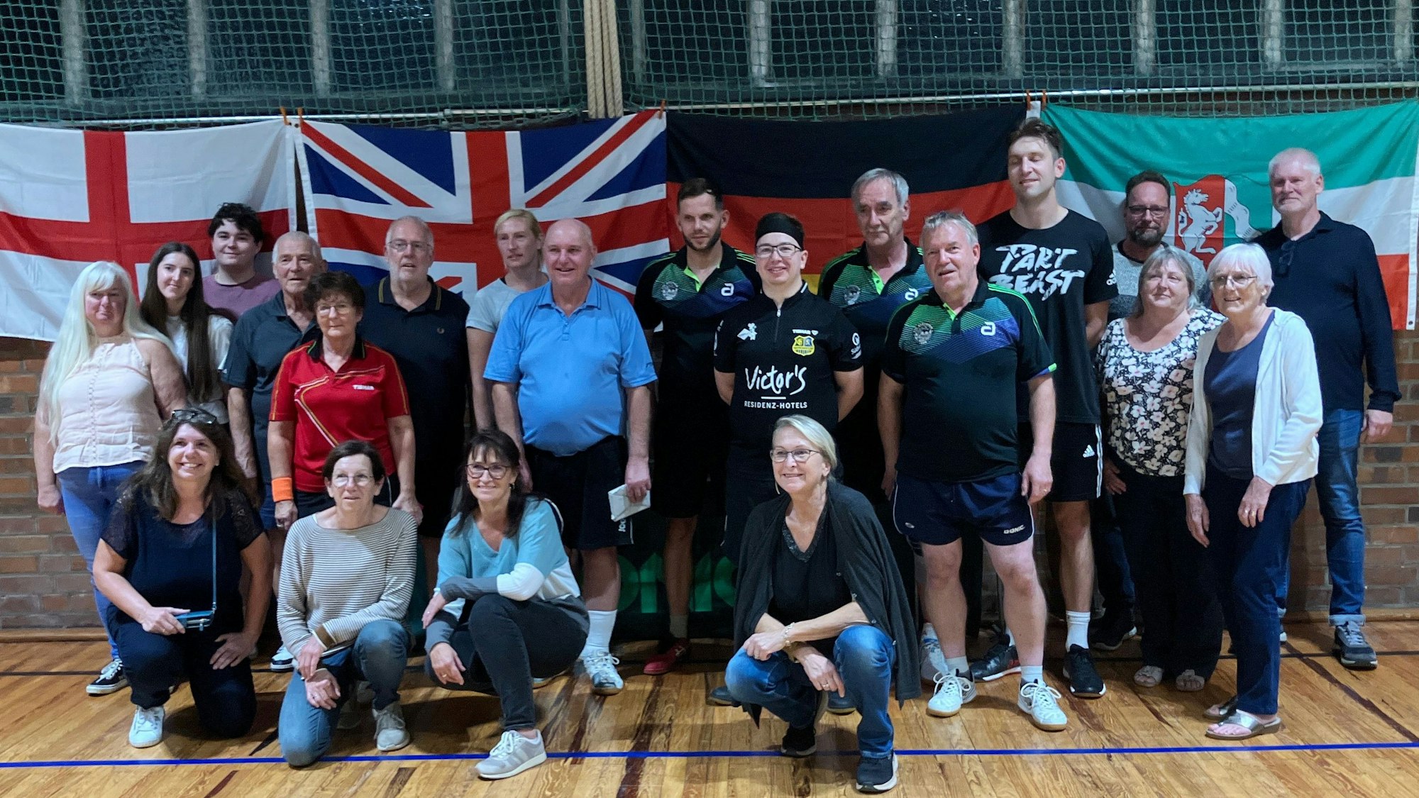 Männer und Frauen stehen in einer Sporthalle, hinter ihnen hängen mehrere Flaggen.