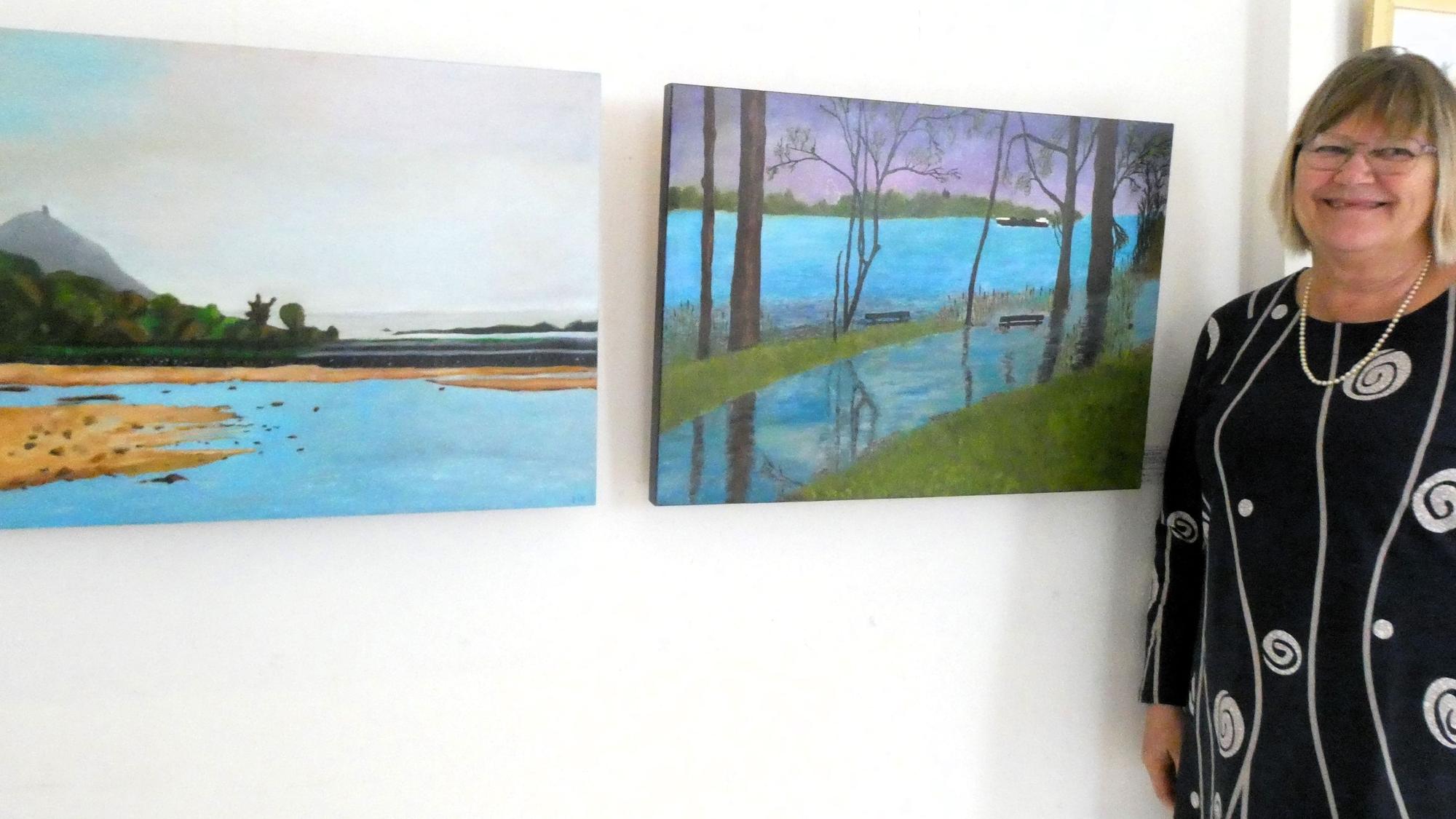 Eine Frau neben zwei Kunstwerken, von denen eines den Rhein und den Drachenfels zeigt.