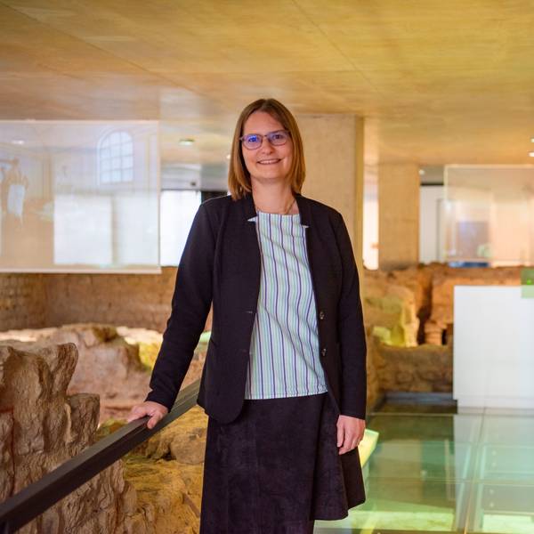 Dr. Silva Bruder lehnt in gestreifter Bluse und dunkelblauem Blazer an einem Geländer in der Ausstellung des Museums für Badekultur. Um sie herum sind die archäologischen Überreste einer antiken Thermen-Anlage zu sehen.