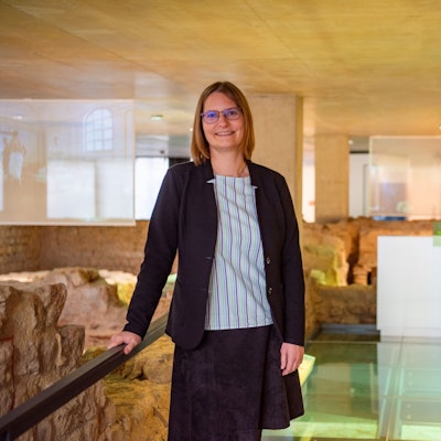 Dr. Silva Bruder lehnt in gestreifter Bluse und dunkelblauem Blazer an einem Geländer in der Ausstellung des Museums für Badekultur. Um sie herum sind die archäologischen Überreste einer antiken Thermen-Anlage zu sehen.