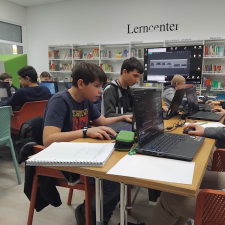 Fünf Jungen im Alter von 13 bis 15 Jahren sitzen vor aufgeklappten Laptops an einem Tisch und schauen konzentriert. Neben ihnen liegen Notizblöcke. Im Hintergrund sieht man weitere Jugendliche und einen großen Bildschirm, außerdem die Bücherregale der Bibliothek. "Lerncenter" steht in schwarzen Lettern an der Wand.