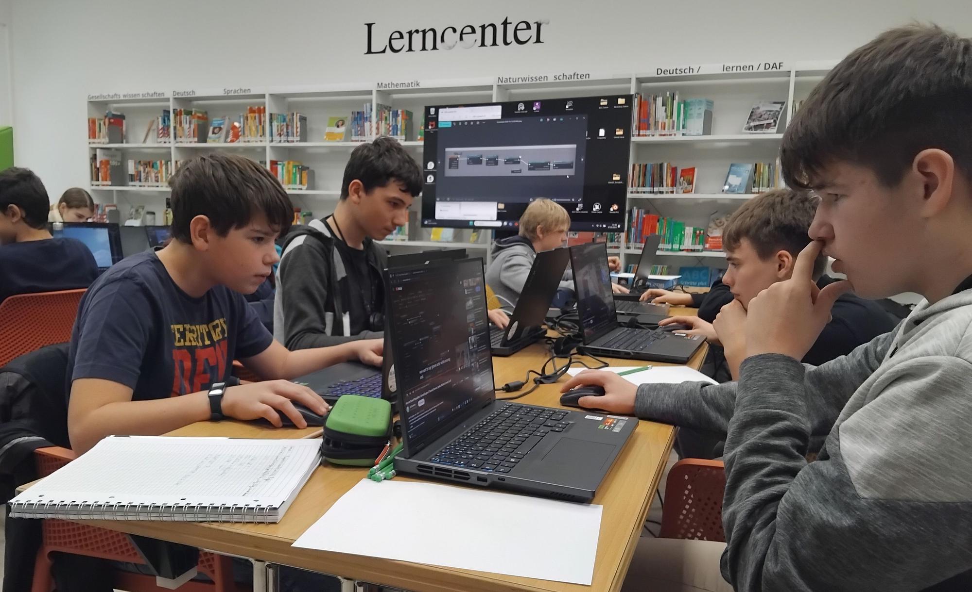 Fünf Jungen im Alter von 13 bis 15 Jahren sitzen vor aufgeklappten Laptops an einem Tisch und schauen konzentriert. Neben ihnen liegen Notizblöcke. Im Hintergrund sieht man weitere Jugendliche und einen großen Bildschirm, außerdem die Bücherregale der Bibliothek. "Lerncenter" steht in schwarzen Lettern an der Wand.