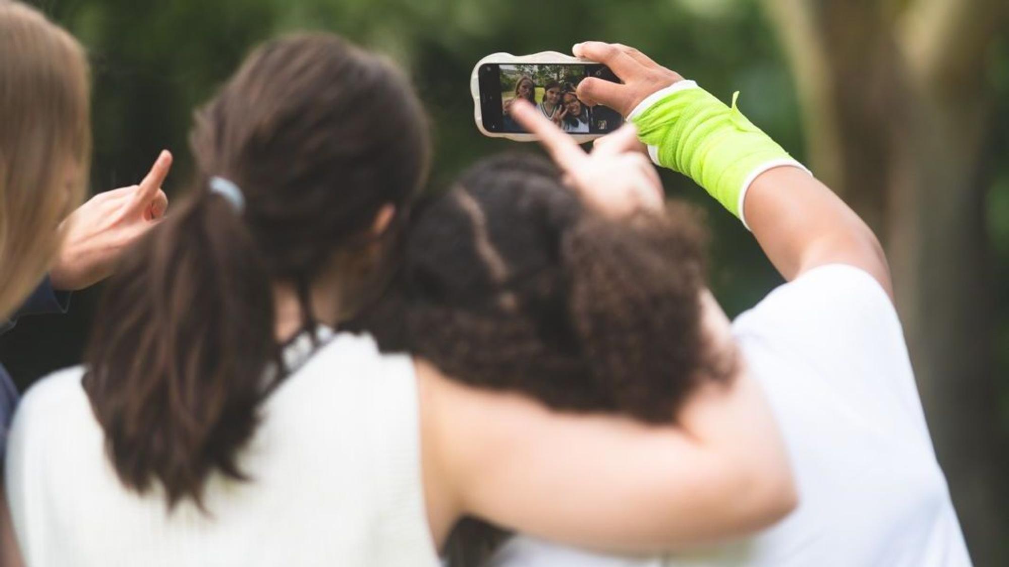 Drei Teenagerinnen sind von hinten zu sehen, sie umarmen sich und machen ein Selfie-Video mit dem Smartphone,