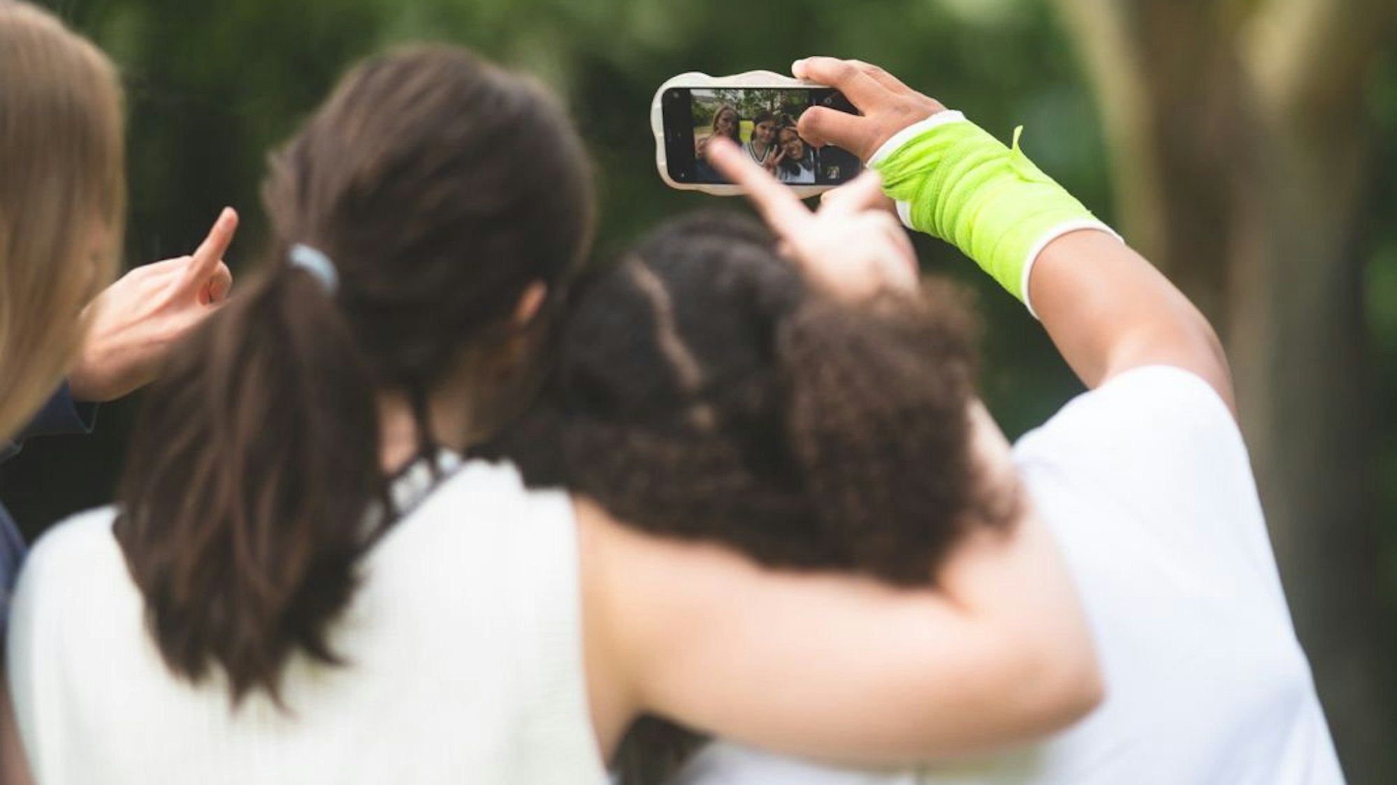 Drei Teenagerinnen sind von hinten zu sehen, sie umarmen sich und machen ein Selfie-Video mit dem Smartphone,