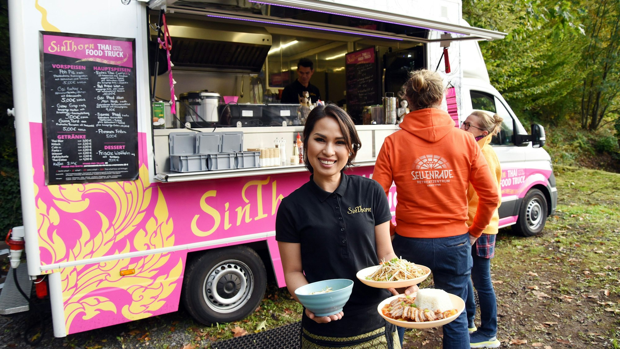 Eine junge Frau steht mit drei Tellern mit thailändischen Gerichten in der Hand vor einem Foodtruck.