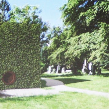 Ein grün bewachsener, großer Kubus mit runder Öffnung steht zwischen alten Grabsteinen und Bäumen.