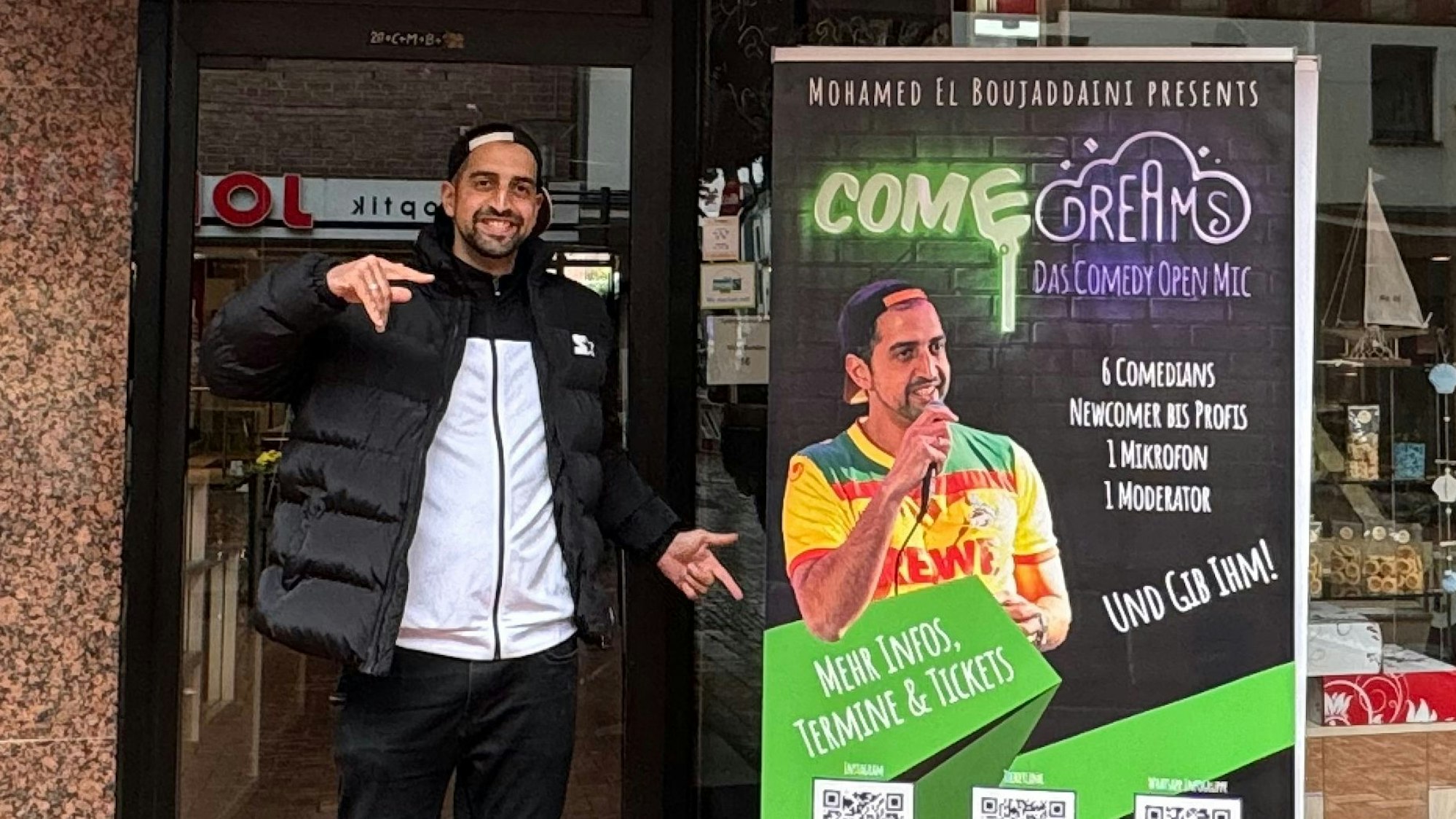 Auf dem Bild ist der Comedian vor einem Veranstaltungsplakat zu sehen.