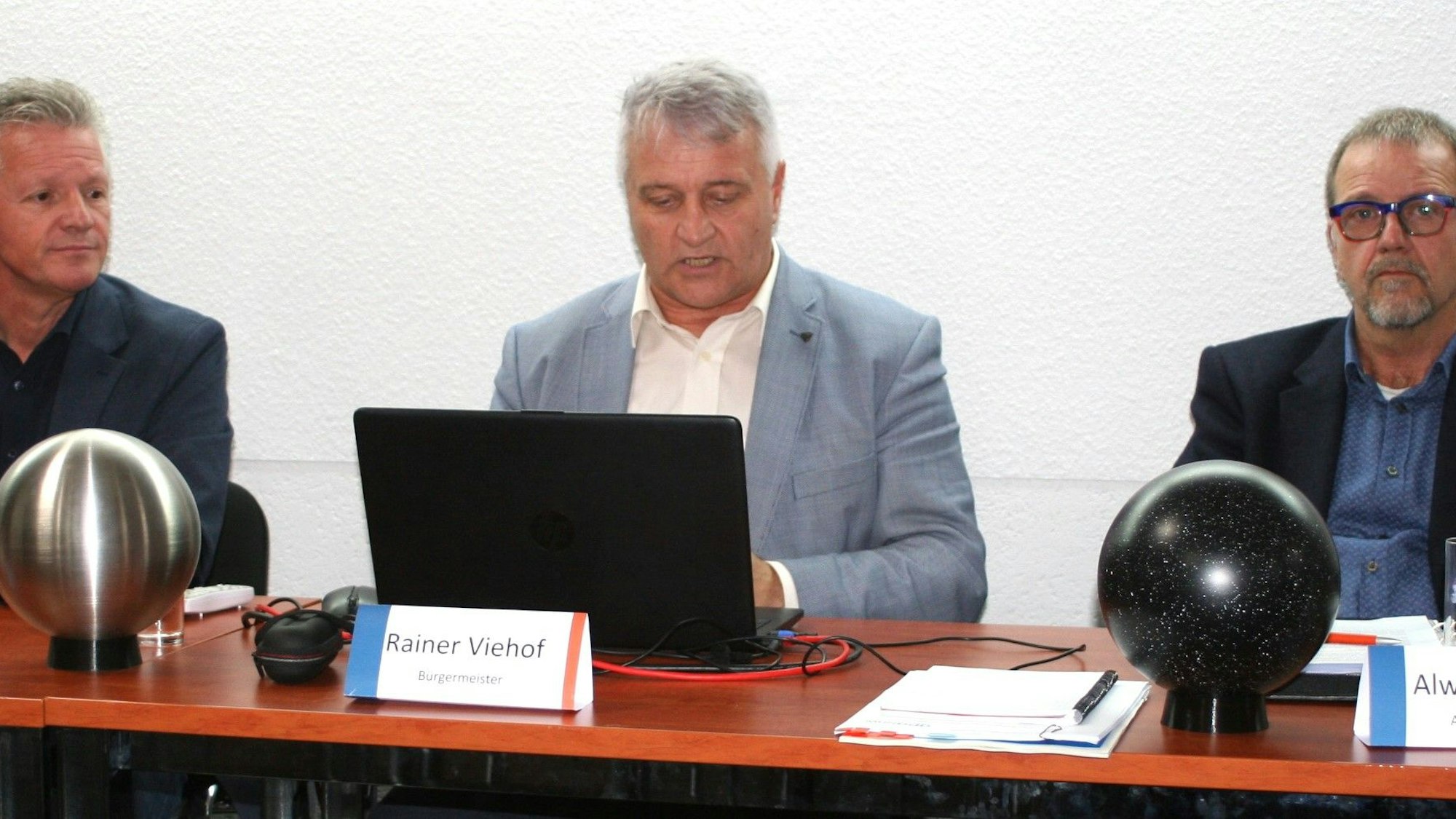 Drei Männer sitzen nebeneinander an einem Tisch, vor ihnen stehen zwei Kugeln.