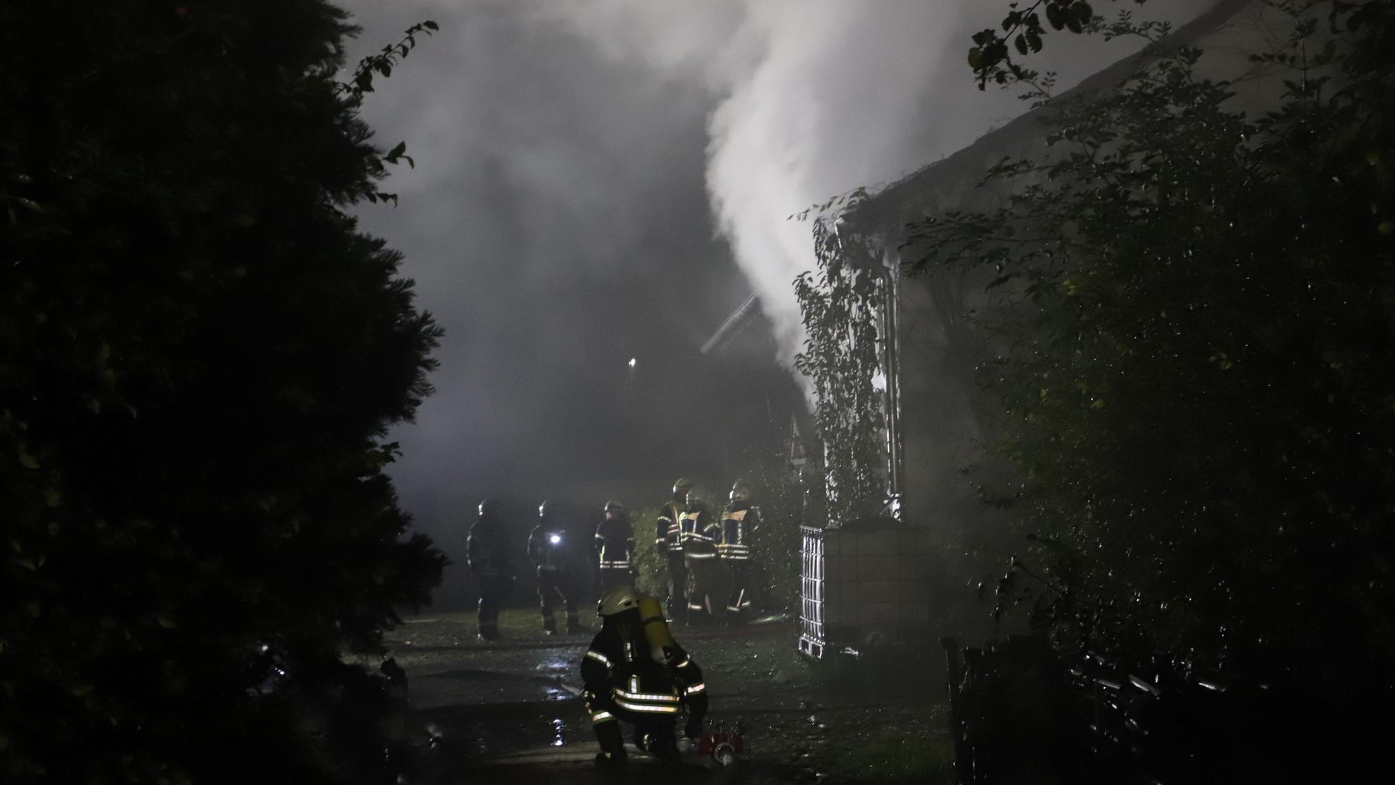 Feuerwehrleute stehen vor einem Haus, aus dem es raucht.